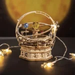 A detailed wooden mechanical model of a solar system with planets, orbit rings, and decorative elements, illuminated by string lights, set against a warm background.