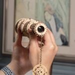 A wooden telescope model held by a person's hand, featuring intricate gears and a chain with a circular charm, designed as a decorative and educational science gadget.