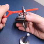 Close-up of a magnetic compass being held and manipulated with a red plastic tool, showing the device's metal needle and circular base on a dark surface.