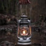 Vintage camping lantern with a small flame, held by a hand over a natural stream in a forest setting, creating a cozy and nostalgic atmosphere.