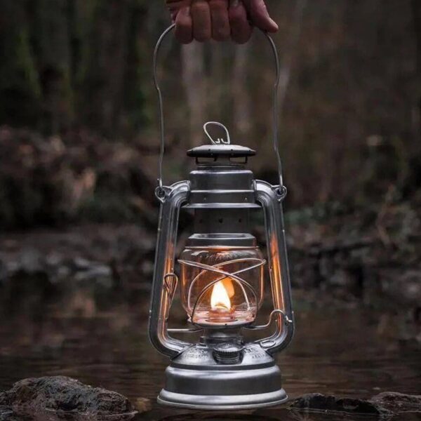 Vintage camping lantern with a small flame, held by a hand over a natural stream in a forest setting, creating a cozy and nostalgic atmosphere.