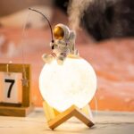 Astronaut figurine sitting on a glowing moon-shaped lamp, creating a space-themed decorative piece. Perfect for science-inspired decor and educational toy displays.