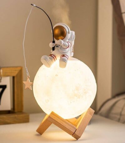 Astronaut figurine sitting on a moon-shaped lamp, holding a fishing rod with a star, creating a whimsical space scene.