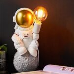 Cute astronaut figurine holding a glowing light bulb, sitting on a moon-shaped base, perfect for science decor and educational spaces.