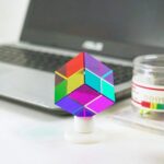 A vibrant, multicolored magnetic puzzle cube with reflective surfaces, placed on a white stand in front of a laptop and a small container of colored magnets, illustrating a science-themed educational.