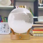 Modern magnetic levitation lava lamp with floating wax globules in a clear glass sphere, set on a wooden base, in a cozy study room.