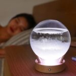 A glowing crystal ball-shaped science lamp on a wooden base with a sleeping person in the background.