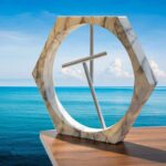 Modern hexagonal marble clock with minimalist metal hands, placed outdoors on a wooden surface overlooking the ocean.