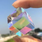 Hand holding a transparent, colorful crystal prism with rainbow reflections against a blue sky background.