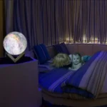 A glowing moon lamp on a stand illuminates a cozy bedroom with two children playing on a bed, surrounded by soft lighting and curtains, creating a warm and educational atmosphere.