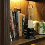 A magnetic levitation science toy displayed on a wooden bookshelf, featuring a clear glass tube with floating particles held above a black base, surrounded by books and decorative items.