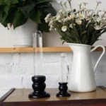 Two clear glass vortex science containers with black bases on a wooden surface, next to a white pitcher filled with daisies, against a white brick wall background.