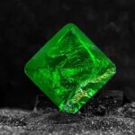 Close-up of a glowing green crystal cube with intricate internal patterns, resting on a textured black surface against a dark background.