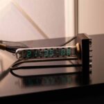 Modern digital clock made from a glass vacuum tube displaying the time 21:35:08, connected to a black power supply with multiple wires, placed on a reflective black surface.