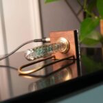Modern digital thermometer with a glass tube, LED display, and wooden frame, placed on a reflective surface with a plant in the background.