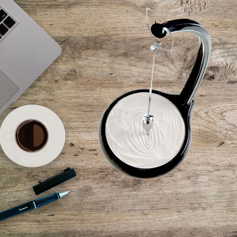 A modern Newton's Cradle with a sleek black and silver design, placed on a wooden desk alongside a cup of coffee, pen, and laptop, illustrating a blend of science and workspace decor.