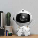 A cute robot camera with a smiling face and starry eyes, sitting on a wooden surface next to a cactus and some small science-related items.