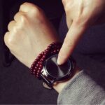 Close-up of a smartwatch on a person's wrist with a finger touching the screen, complemented by red beaded bracelets.
