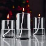 Three glass candles with floating wicks and lit flames, creating a warm glow against a dark background with blurred colorful lights.