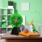 A Tesla coil lamp with a glowing green plasma sphere on a black base, placed on a wooden desk in a modern, colorful room.