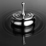 An artistic black and white photograph of a water droplet splash captured mid-air, creating a smooth, reflective surface and ripples in the water below.