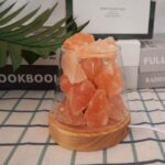 Himalayan salt crystals displayed in a clear glass jar on a wooden base, with a background of books and a plant leaf, showcasing natural mineral decor.