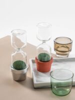 Glass hourglass with green moss in small pots, decorative science-themed items for home or classroom.