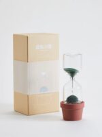 Modern sand timer with black sand flowing through an hourglass, placed in a small terracotta pot, with a plain white background.