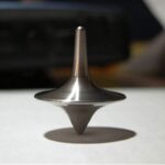 Metal spinning top toy on a white surface, casting a shadow, with a blurred background.
