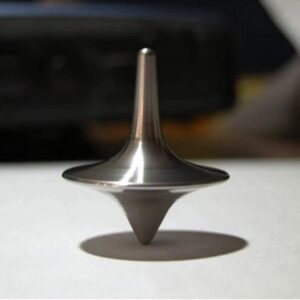 Metal spinning top toy on a white surface, casting a shadow, with a blurred background.