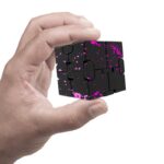 Hand holding a black puzzle cube with pink paint splatter pattern, showcasing a challenging and colorful brain teaser toy for educational and recreational use.