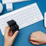 Hand holding black cube puzzle over a white keyboard on a blue desk, with science-themed decor and educational toys in the background.