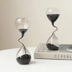 Two hourglass-shaped glass timers with black sand, placed on a white surface, one on a book, showcasing science decor and educational toys.