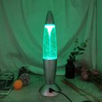 Modern lava lamp emitting a green glow with swirling wax inside, placed on a desk with decorative items and plants in the background.