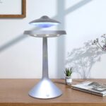 Modern UFO-shaped LED lamp with blue light on a wooden desk, accompanied by a potted plant and a magazine, creating a sleek and futuristic decor element.