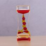 Transparent hourglass with red and yellow beads flowing from top to bottom, demonstrating the passage of time in a clear, visually appealing design.