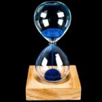 Elegant glass sand timer with blue sand flowing between two chambers, mounted on a wooden base, ideal for educational and decorative purposes.