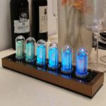 Modern nixie tube clock displaying glowing blue and green numbers, placed on a white surface with wine bottles and glasses in the background.