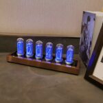 Modern nixie tube clock displaying the time with blue LED lights, placed on a wooden base, next to framed artwork, on a gray surface.
