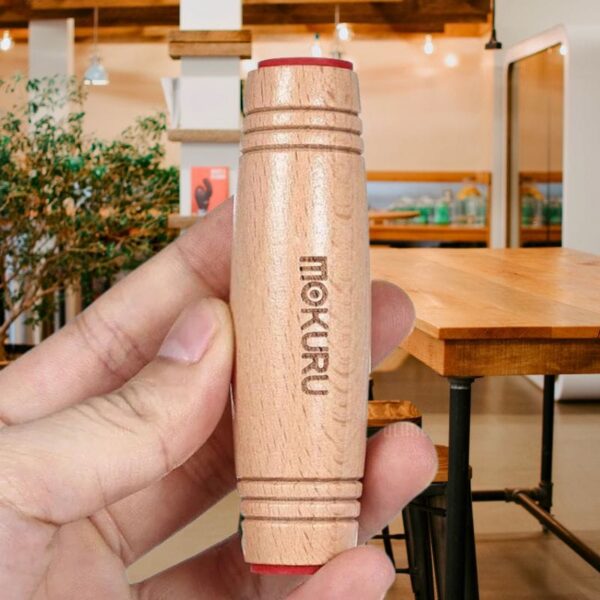 A close-up of a wooden educational toy labeled "MOKURU," held by a person's hand inside a cozy, well-lit store with wooden furniture and decor.