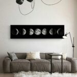 A sleek black wall art piece displaying the different phases of the moon, mounted above a contemporary gray sofa in a minimalist living room.