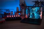 Nixie tube clock with glowing numerals and a laptop on a desk, surrounded by science-themed decor and toys, illuminated by blue ambient lighting.
