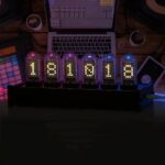 Modern digital nixie tube clock displaying the time with vibrant, colorful tubes on a desk setup, surrounded by a laptop, MIDI controller, and other tech accessories.