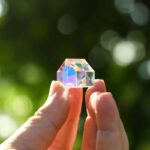 Close-up of a glass prism held between fingers, displaying rainbow reflections and light refraction against a blurred green outdoor background.