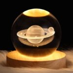 A detailed glass sphere containing a miniature model of a planet with rings, illuminated to highlight its features, placed on a wooden base against a dark background.