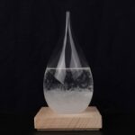 Clear glass oil dropper with a teardrop shape, filled with liquid, mounted on a light wooden base, set against a black background.