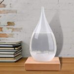 A sleek, water drop-shaped diffuser filled with clear water, placed on a wooden base against a white brick wall.