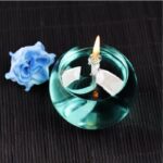 Elegant glass oil lamp with a lit wick, accompanied by a delicate blue rose, placed on a dark textured surface for a sophisticated decorative display.