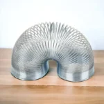 A metallic coil sculpture shaped like a flexible, curved tube, showcasing intricate spirals and a shiny surface, placed on a wooden surface.