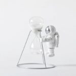 Astronaut figure holding a large hourglass with white sand, standing on a minimalist metal stand.